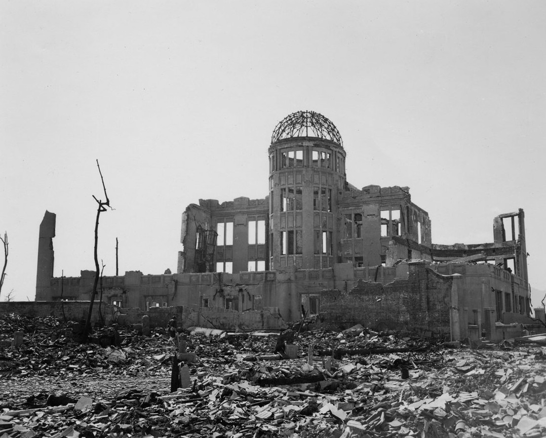 A photo showing Hiroshima Prefectural Commercial Exhibition Hall after the blast. The exterior walls have all been destroyed along with the roof and upper levels. All that remains is the foundation walls of the ground floor and the skeletal frame of the cylindrical structure holding up the dome, which itself has been reduced to a wire frame. The sounding area is covered in layers of rubble and the trees in front of the building are nothing more than chard sticks. A photo showing Hiroshima Prefectural Commercial Exhibition Hall after the blast. The exterior walls have all been destroyed along with the roof and upper levels. All that remains is the foundation walls of the ground floor and the skeletal frame of the cylindrical structure holding up the dome, which itself has been reduced to a wire frame. The sounding area is covered in layers of rubble and the trees in front of the building are nothing more than chard sticks.