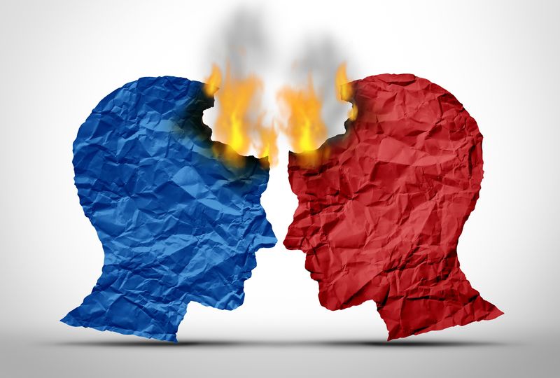 An image showing two male heads on profile that are made of paper. They are facing one another as if in conflict. The one on the left is blue while the one on the right is red. Both paper heads are on fire. 