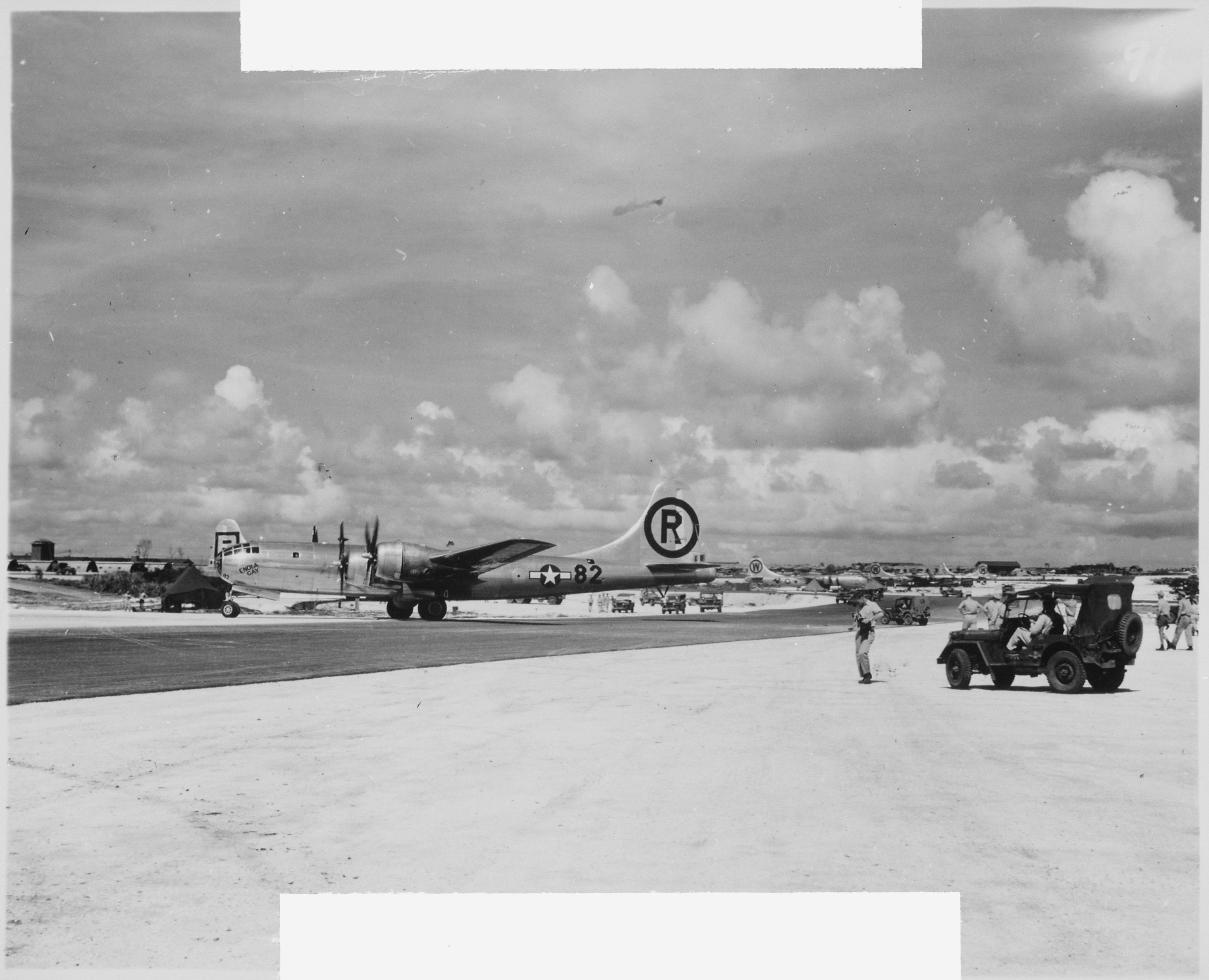 A black and white photo showing the Enola Gay on a runway. The aircraft is shown on profile, facing the left of the shot. It has twin predellas on each wings. There is a large R encircled and printed on its tail and the number 82 is printed on its tail, next to a symbol of a star in a circle with a rectangle running through it. The word "Enola Gay" is written at an angle just under the cockpit window. The aircraft is in the middle of the shot. In the righthand side of the foreground is a jeep with several military personnel standing around it. A black and white photo showing the Enola Gay on a runway. The aircraft is shown on profile, facing the left of the shot. It has twin predellas on each wings. There is a large R encircled and printed on its tail and the number 82 is printed on its tail, next to a symbol of a star in a circle with a rectangle running through it. The word "Enola Gay" is written at an angle just under the cockpit window. The aircraft is in the middle of the shot. In the righthand side of the foreground is a jeep with several military personnel standing around it.