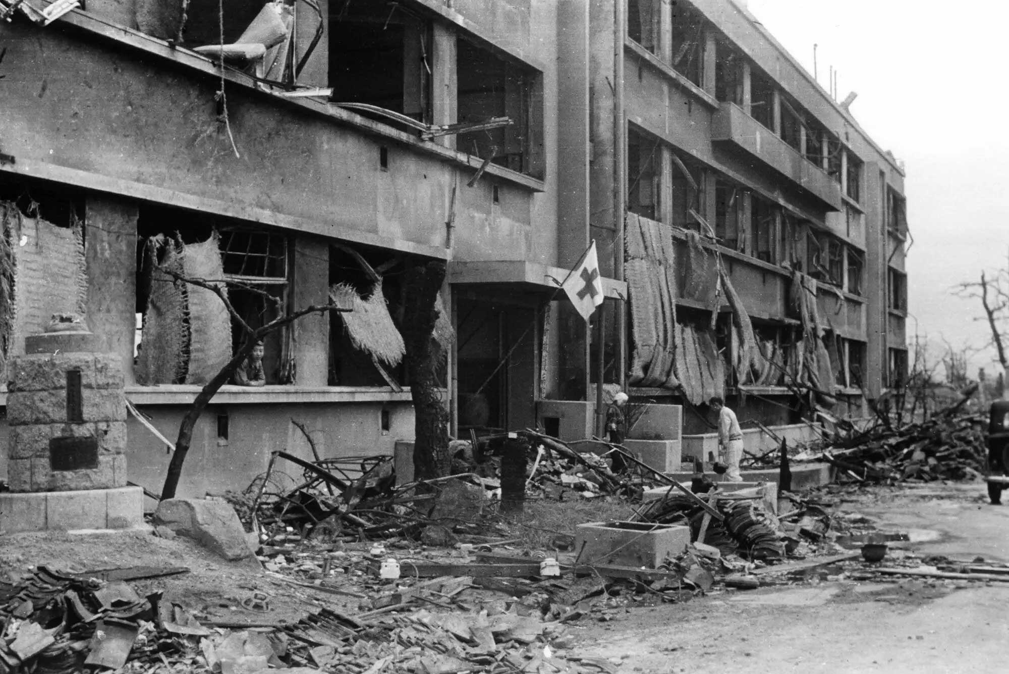 The photo shows a lopsided Red Cross flag attached to the entrance of an otherwise blown out three storied building. The windows are all missing and the front of the building is strewn with rubble and twisted metal. There is a man shown walking towards the entrance. The photo shows a lopsided Red Cross flag attached to the entrance of an otherwise blown out three storied building. The windows are all missing and the front of the building is strewn with rubble and twisted metal. There is a man shown walking towards the entrance.