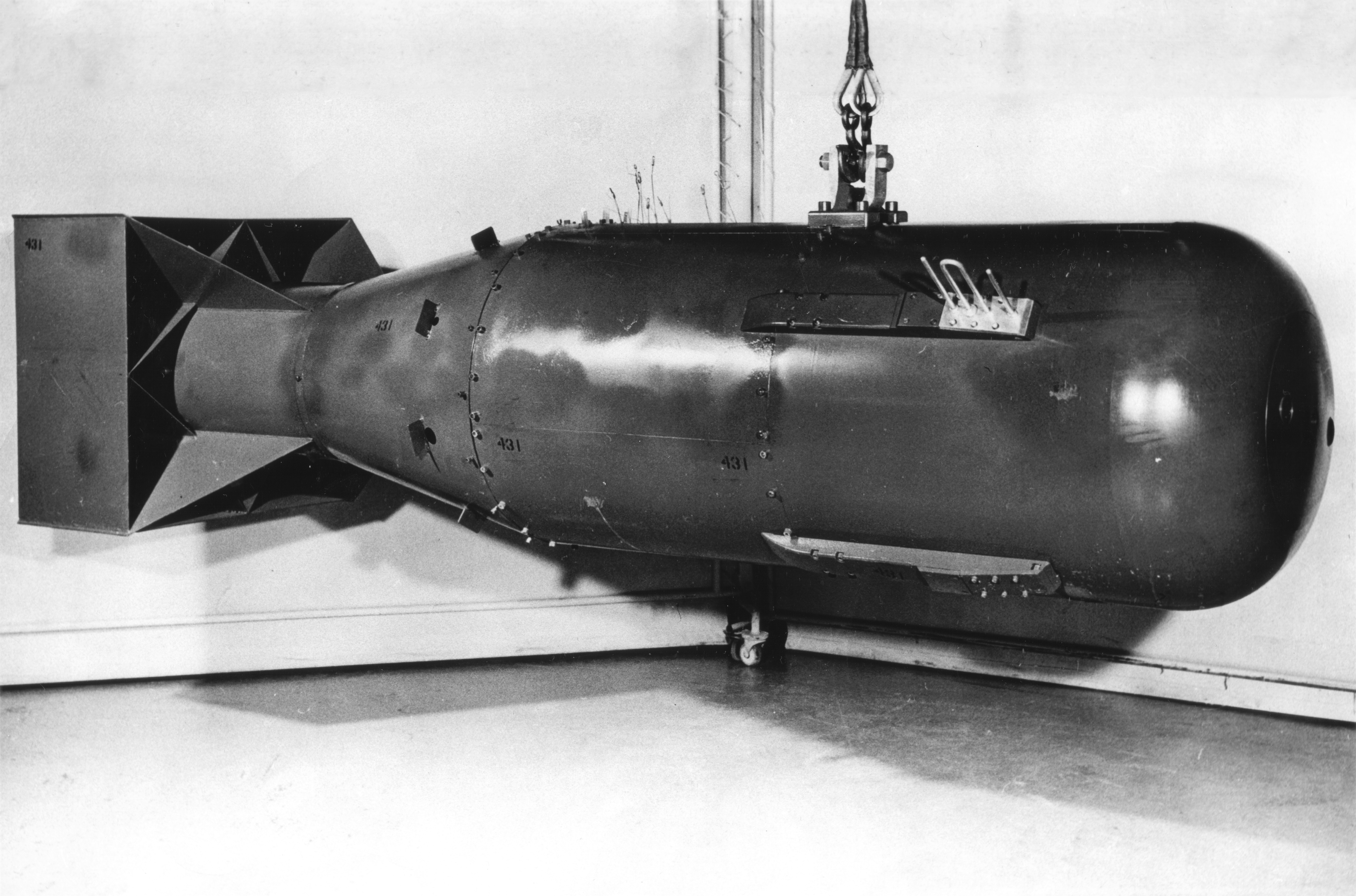 A photo showing the Little Boy bomb on profile. It has the classic bomb shape and appears in dark steel. It is being suspended in the air by a metal cable. A photo showing the Little Boy bomb on profile. It has the classic bomb shape and appears in dark steel. It is being suspended in the air by a metal cable.